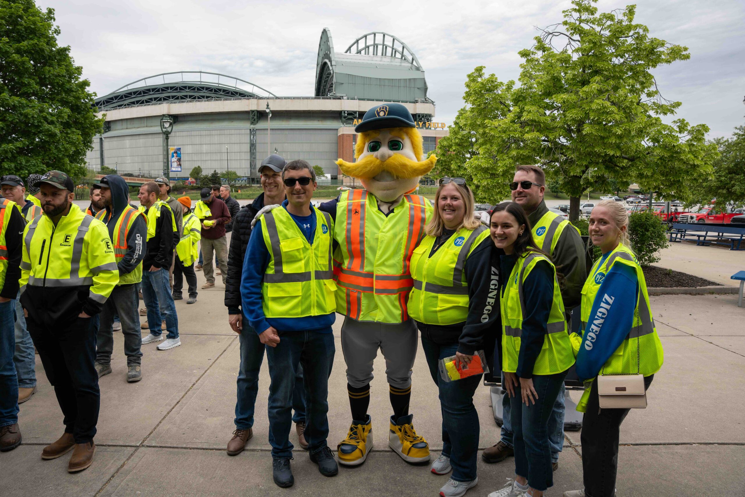 Group of people in bright safety vests posing for a photo outside a stadium, with a mascot wearing a blue cap in the center.