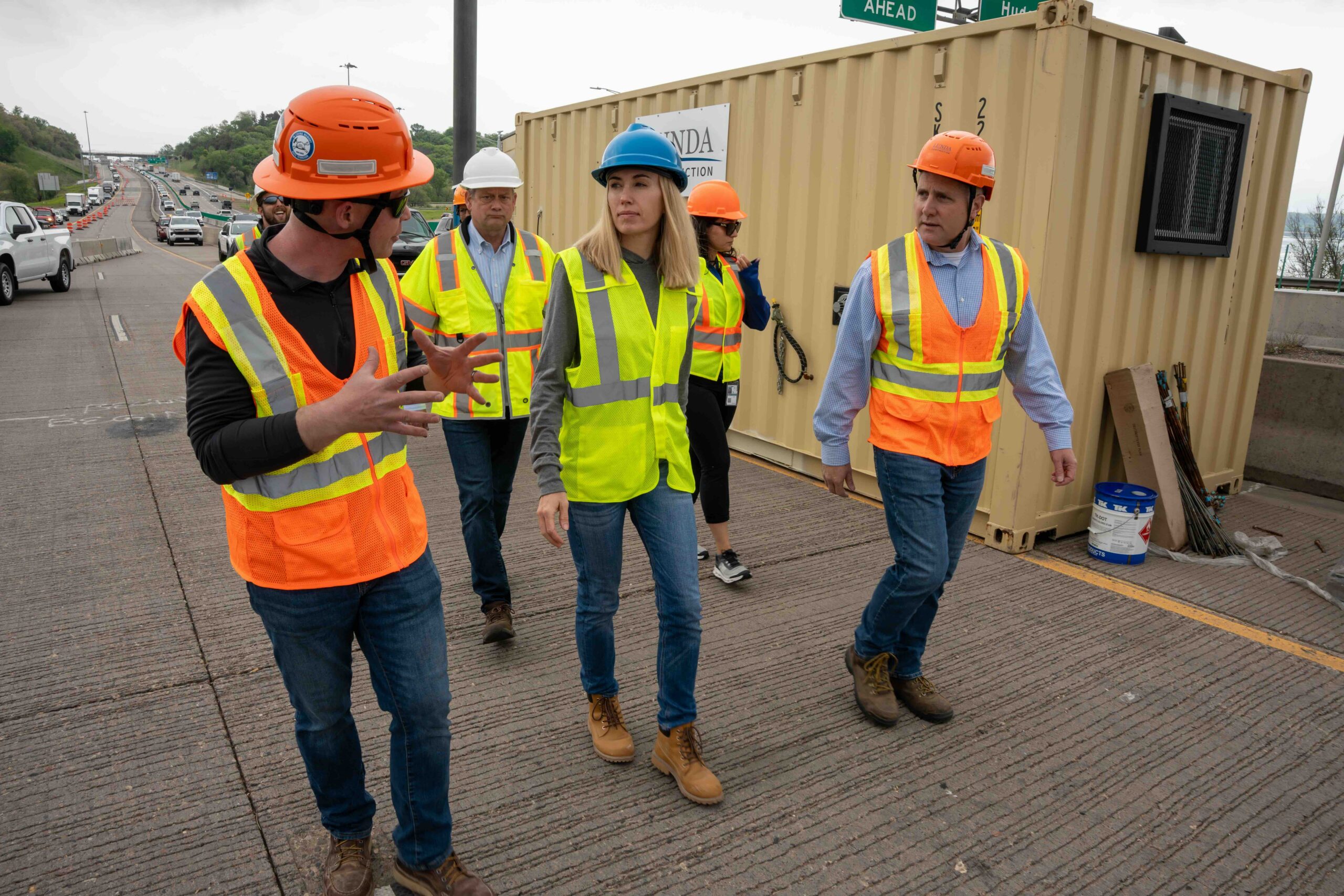 Construction crew in hi-vis vests and hard hats stand near a beige container on a highway worksite.