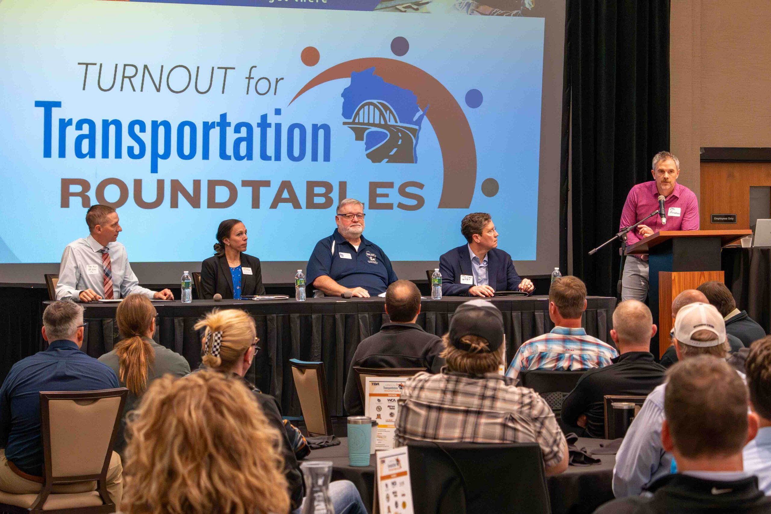 Panel of five people seated at a long table on a stage, with a large banner reading 'Turnout for Transportation Roundtables' behind them; a man at a podium speaks on the right.