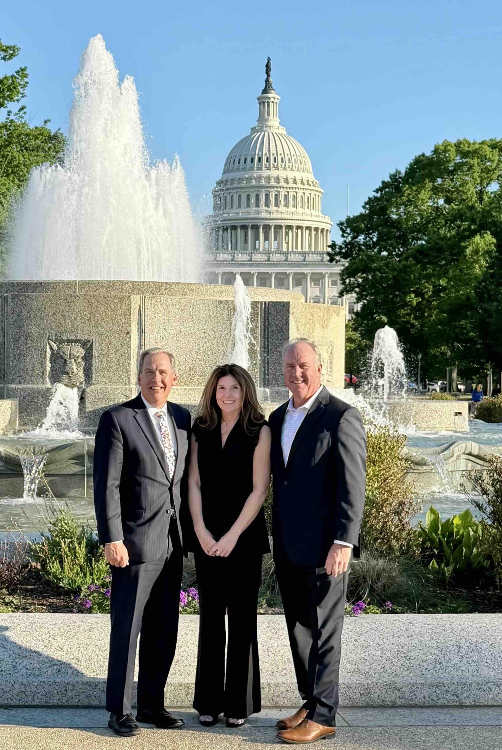 Three professionally dressed adults posing in front of a large fountain with the U.S. Capitol dome in the background on a sunny day.