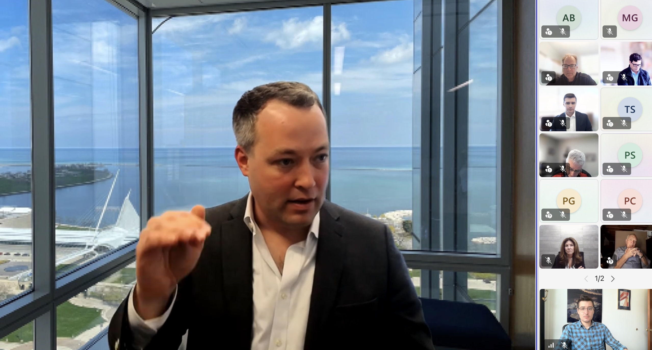 Man in a suit speaks and gestures during a video conference in a glass-walled office with an ocean view outside.