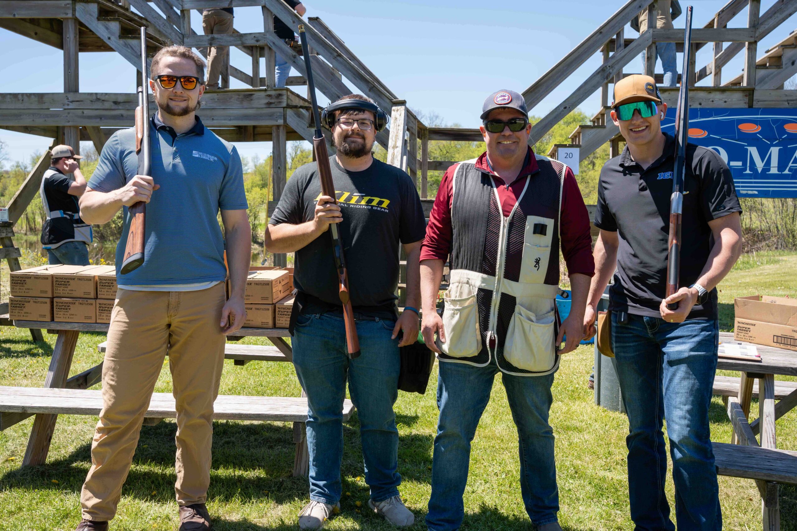 Four men stand outdoors at a shooting range, each holding a rifle, with wooden stands and ammo boxes nearby.