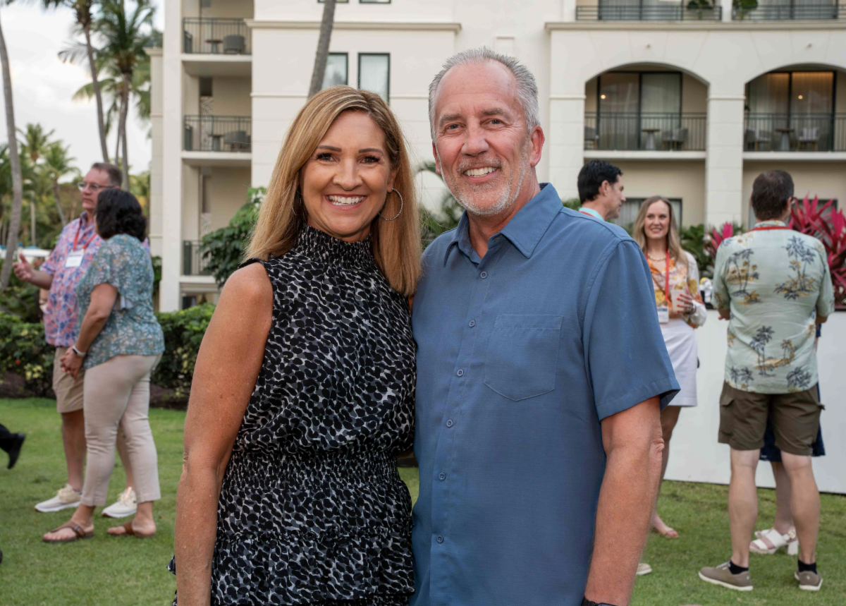 Smiling couple posing outdoors at a casual event, with others mingling and a beige building behind them.