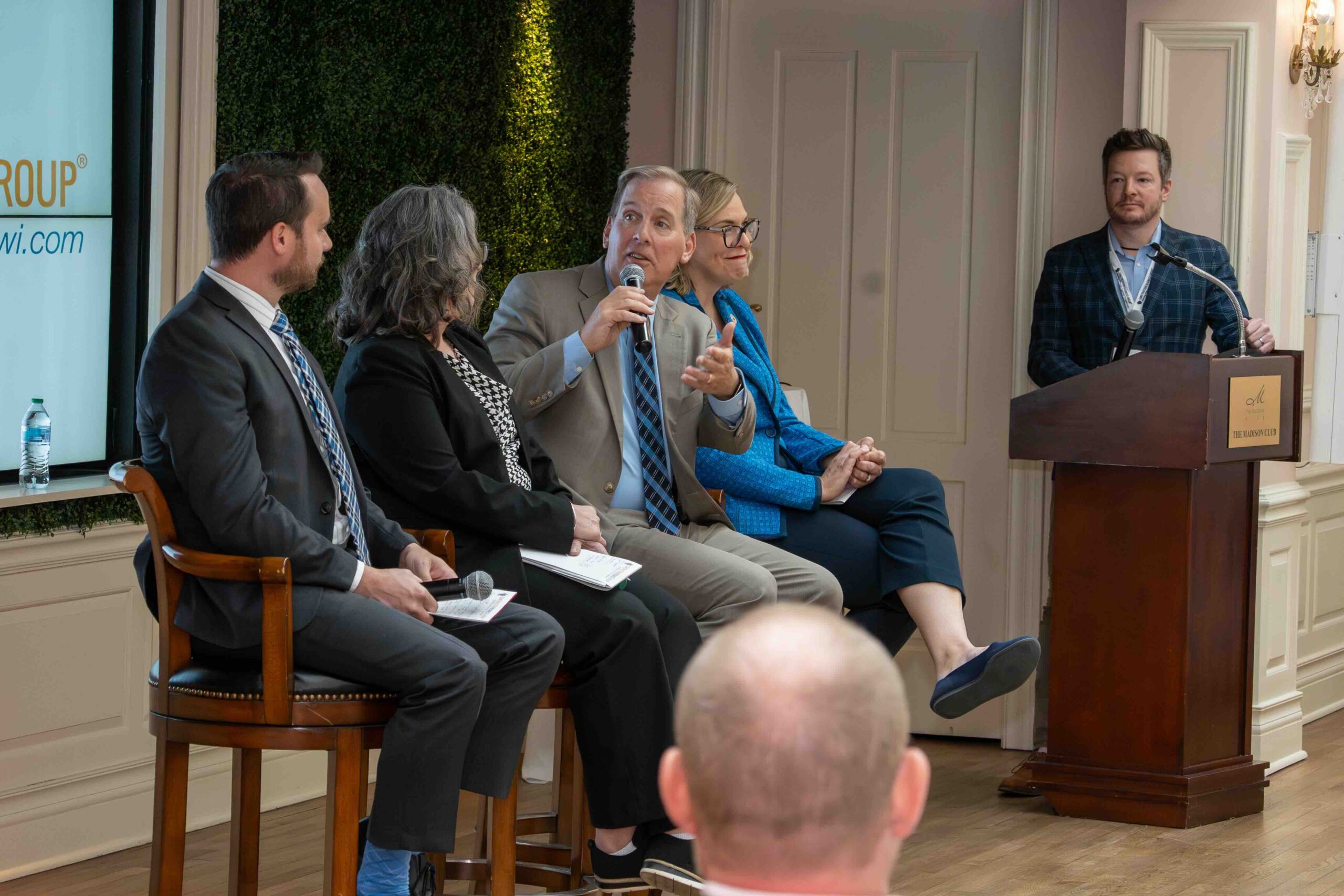 Panel discussion with five people on stage; a man in a beige suit speaks into a handheld microphone while others listen, and a podium is on the right.