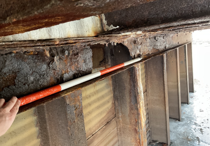 Rusty metal beam with corroded surfaces; a red-and-white striped pole is resting along it, held by a hand on the left.