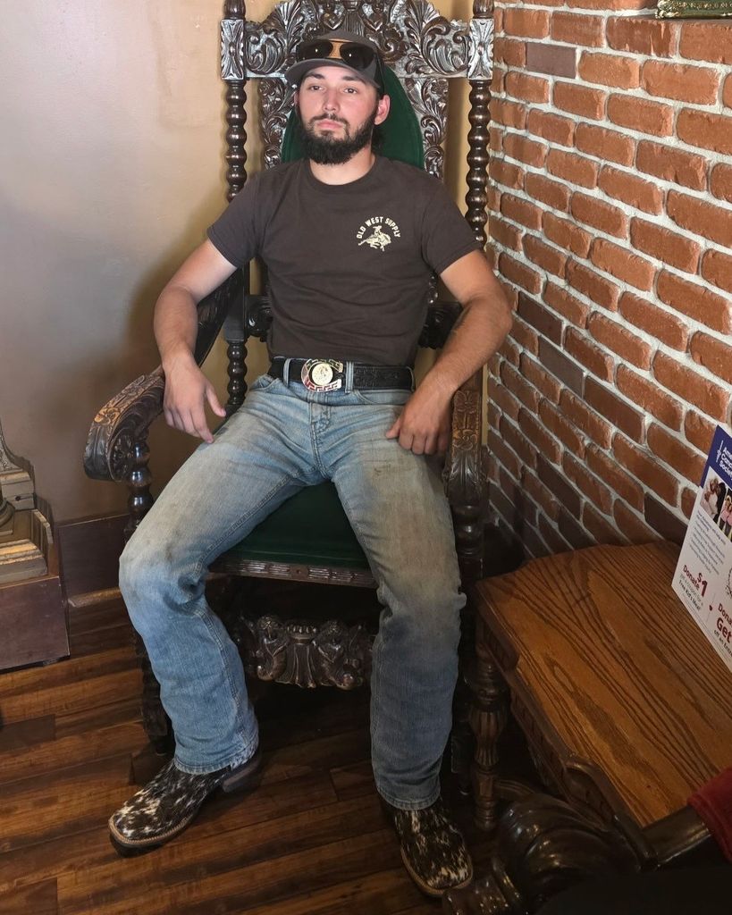 Man with beard sits on an ornate carved throne, wearing a brown t‑shirt, jeans, cowboy belt buckle, and patterned boots, against a brick wall.
