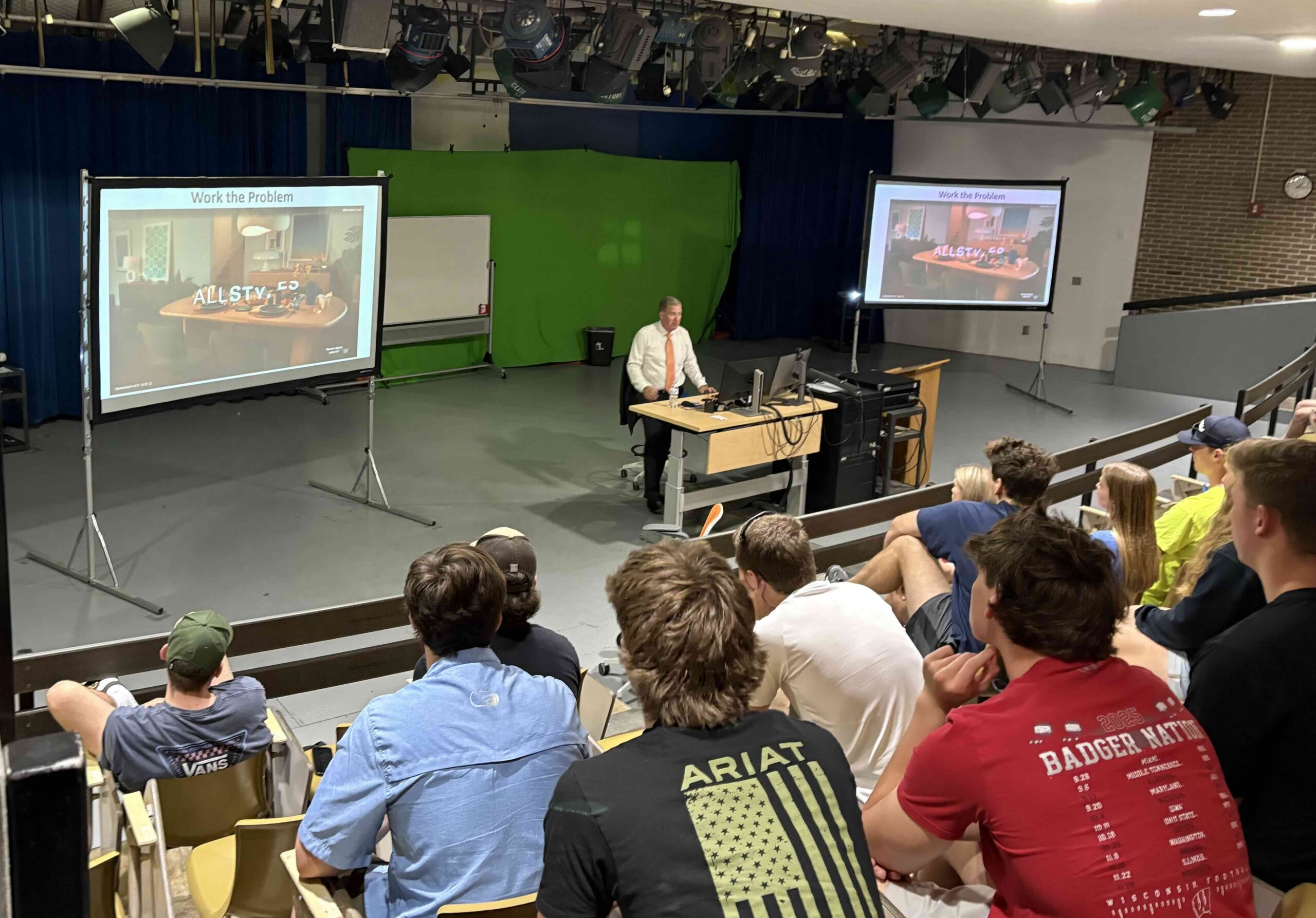 Presenter in a white shirt and orange tie stands at a desk at the front of a lecture hall; two projection screens show slides labeled 'Work the Problem' as students watch.