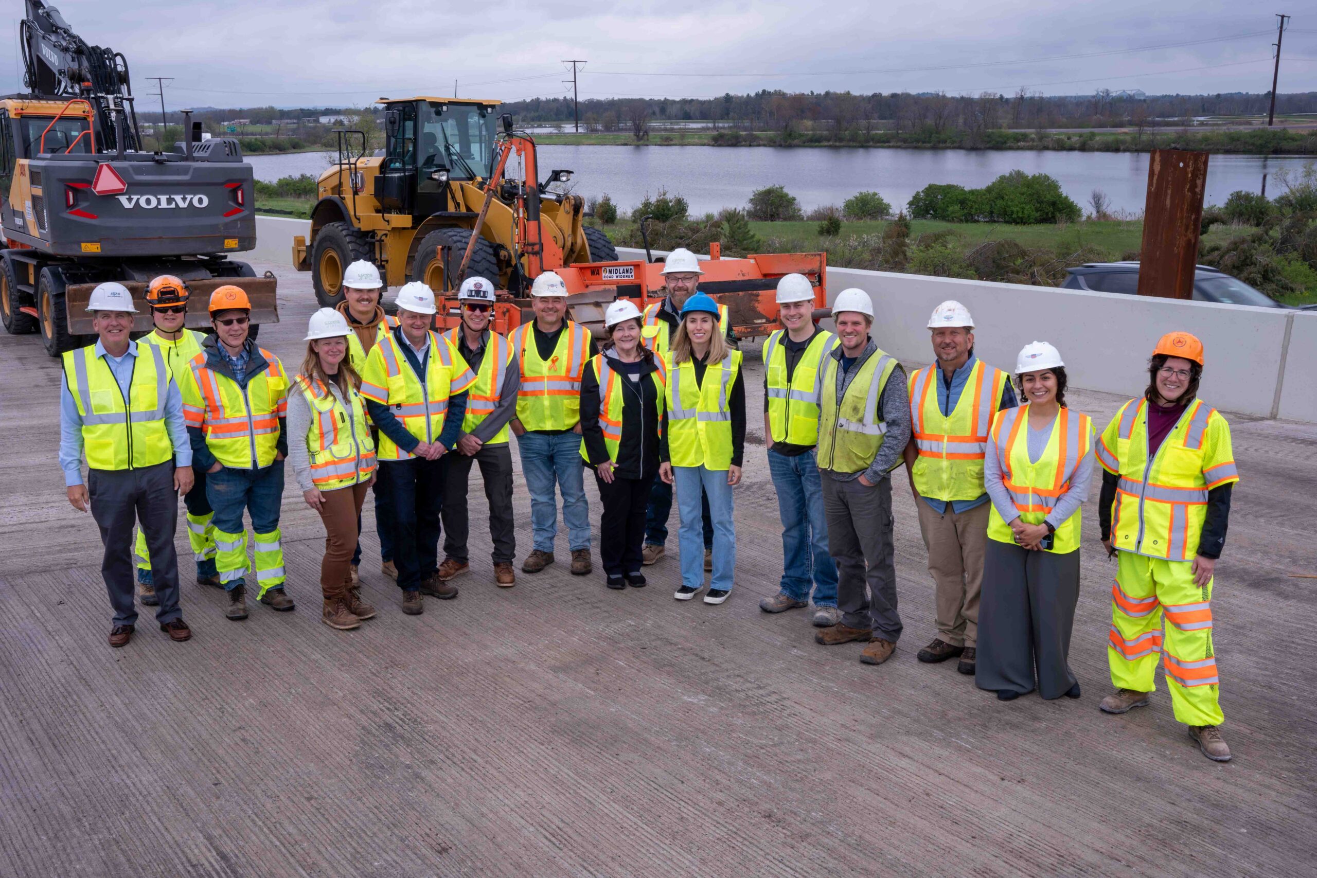 Group of construction workers in safety vests and hard hats posing on a concrete site with heavy machinery in the background and a river beyond.