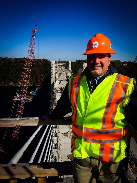 Construction worker in a high-visibility vest and orange hard hat standing on a bridge under construction with a crane in the background.
