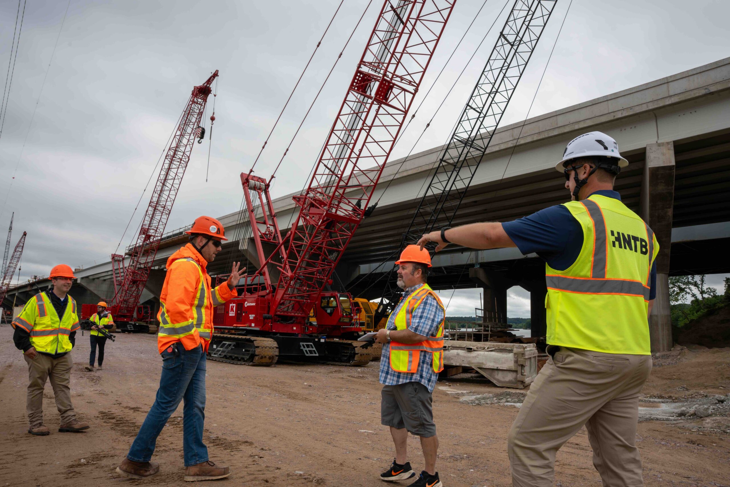 Construction workers in high‑visibility vests and hard hats observe a large red crane at a bridge construction site. (Informative)
