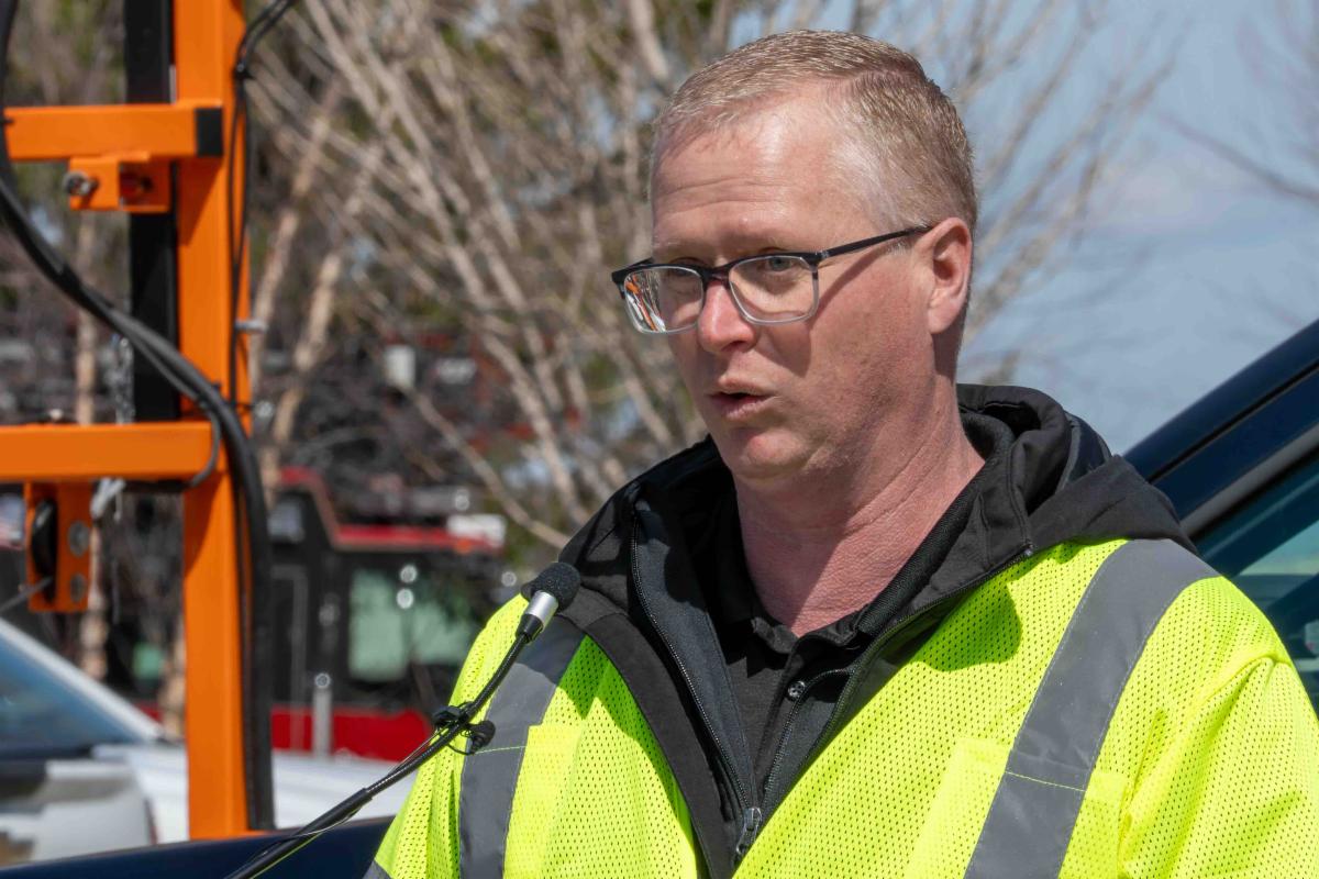 Man in a bright yellow safety vest speaks into a microphone outdoors, likely giving a briefing or interview.
