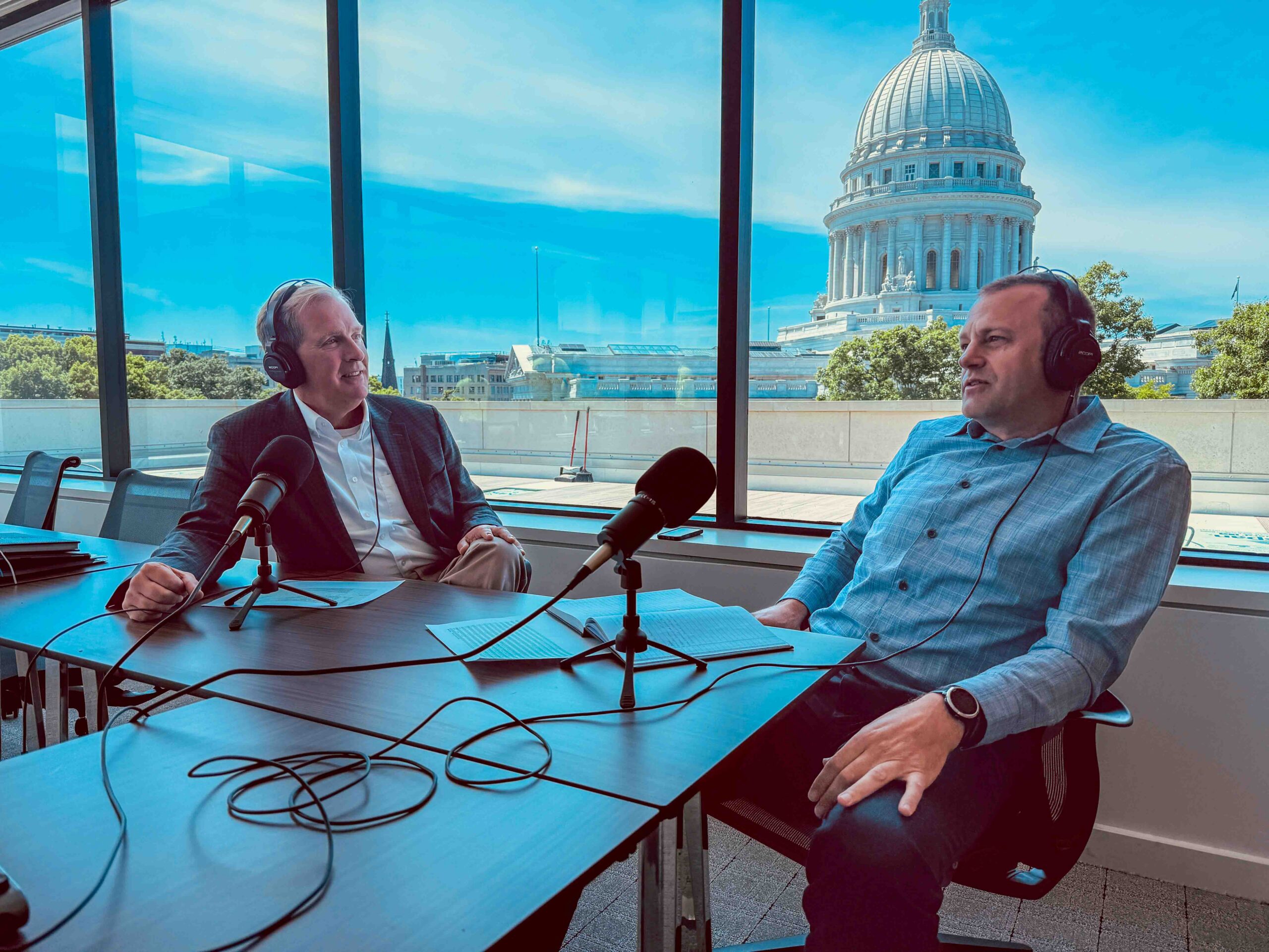 Two men wearing headphones speak into microphones in a glass-walled podcast studio, with a domed government building visible through the windows outside.