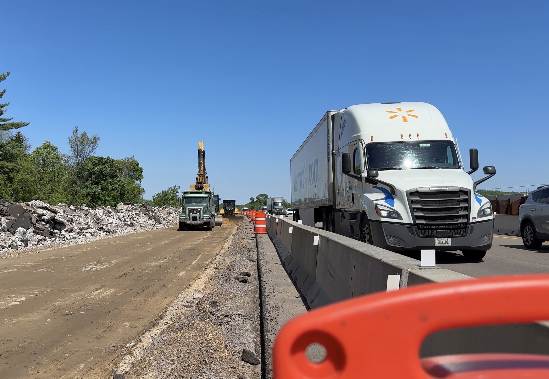 Construction site on a highway: a white semi-truck is parked along a concrete barrier, with orange traffic barrels, a dirt road to the left, and rock piles under a clear blue sky.