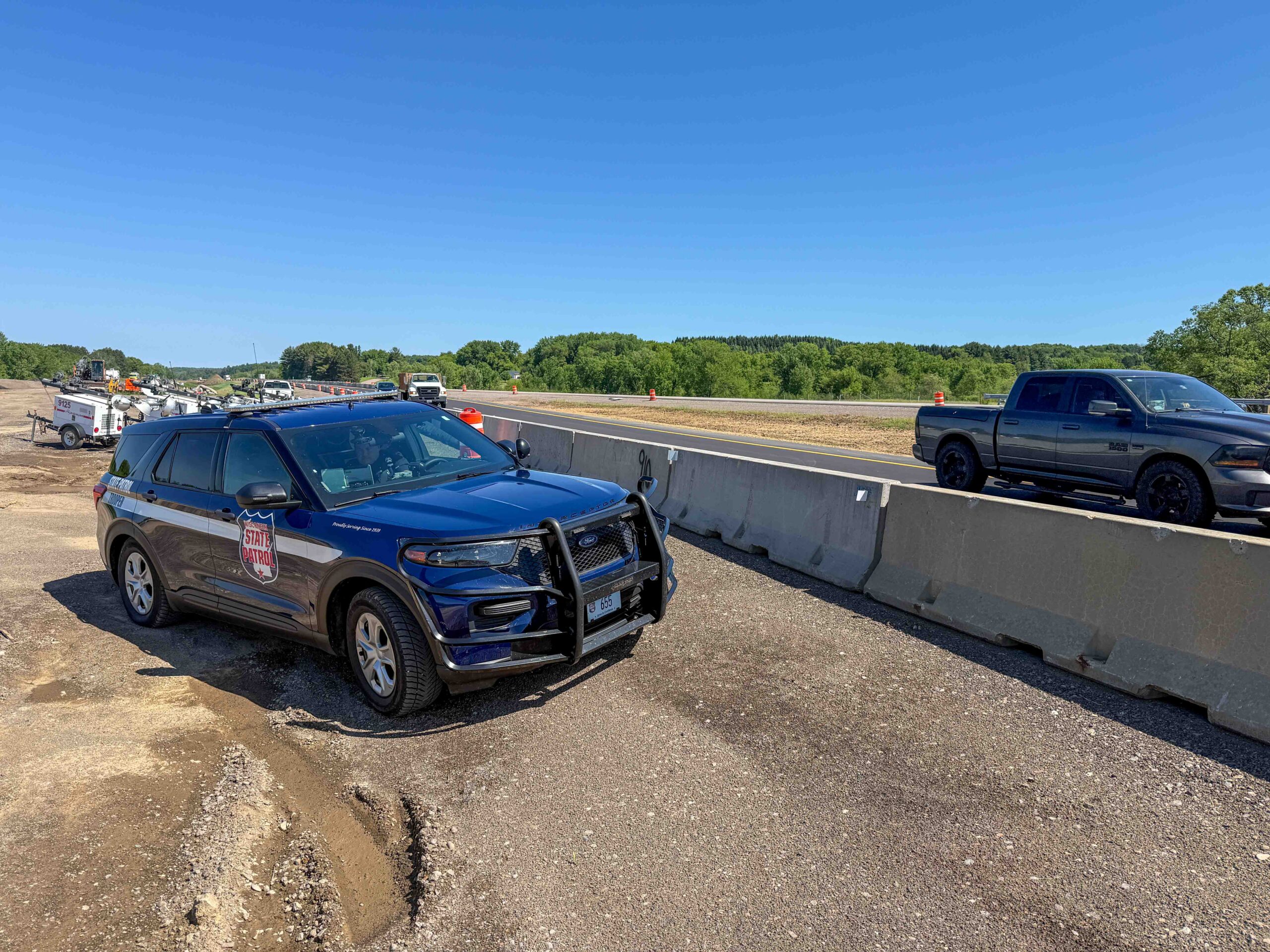 State Patrol SUV in the foreground at a highway construction site, with concrete barriers, orange traffic barrels, and distant construction equipment under a clear blue sky.