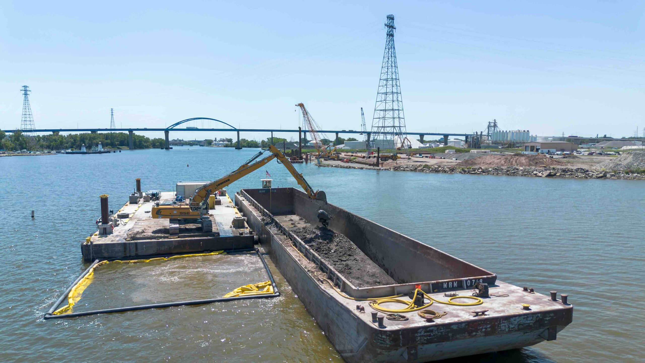Construction barge on a river with an excavator digging soil, cranes, and a bridge in the distance, under a clear blue sky.