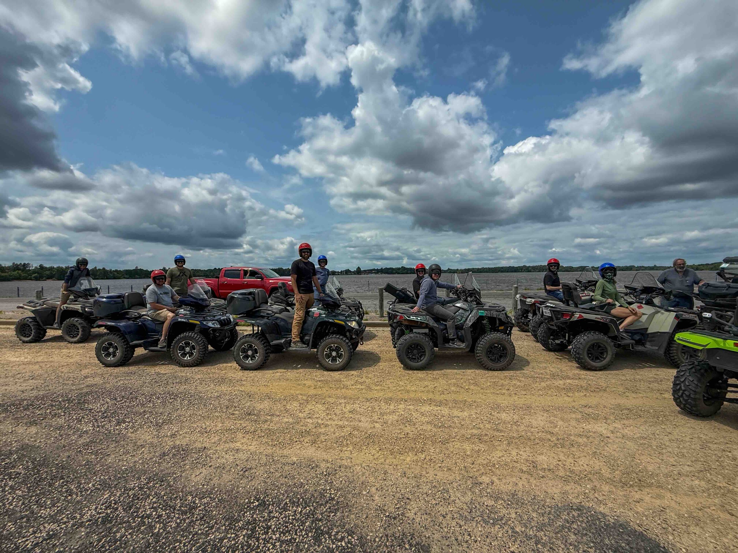 Group of people wearing helmets beside parked ATVs along a lakeside under a cloudy sky.