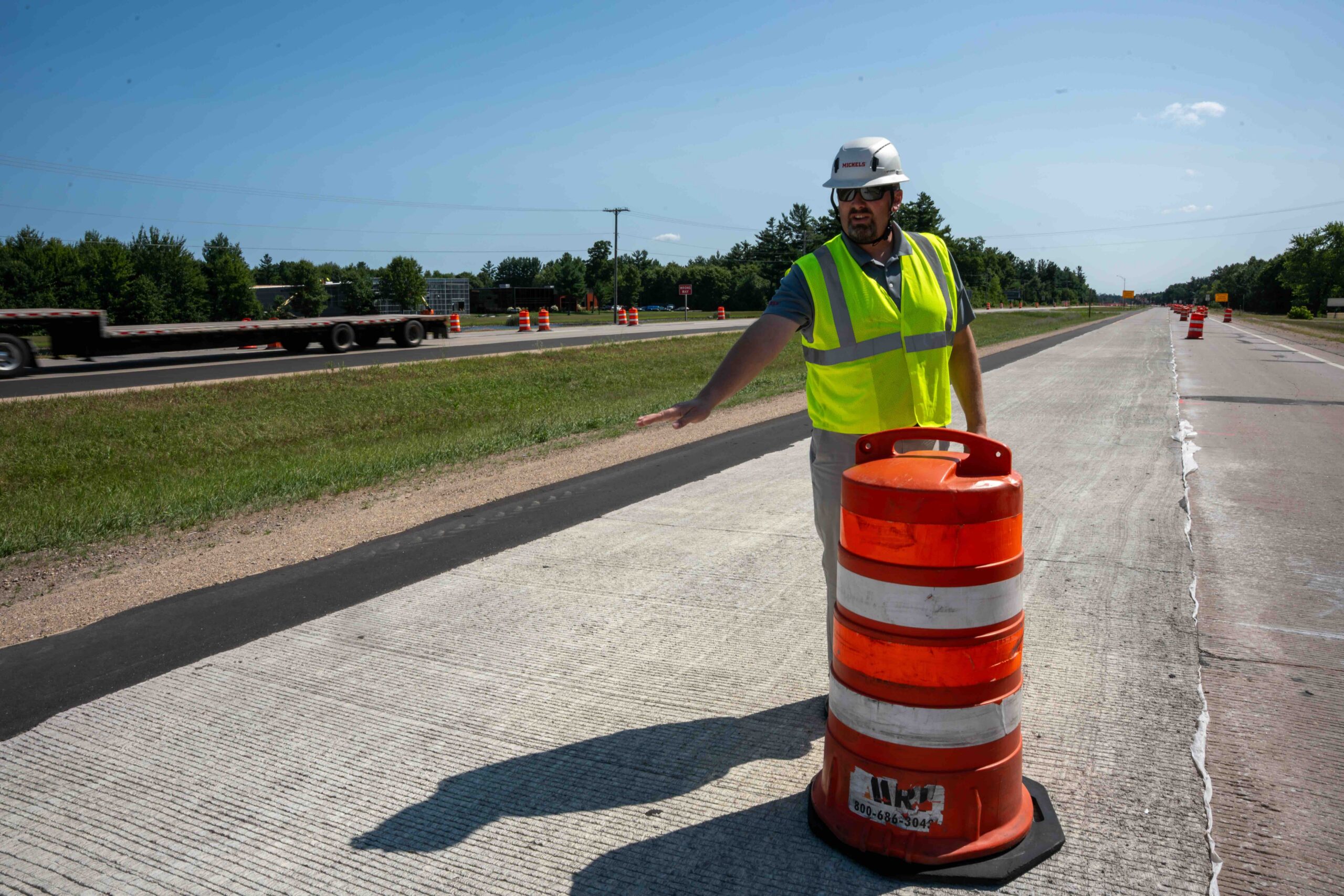 Construction worker in a neon safety vest and white hard hat stands by orange traffic barrels directing traffic on a highway under construction on a sunny day.