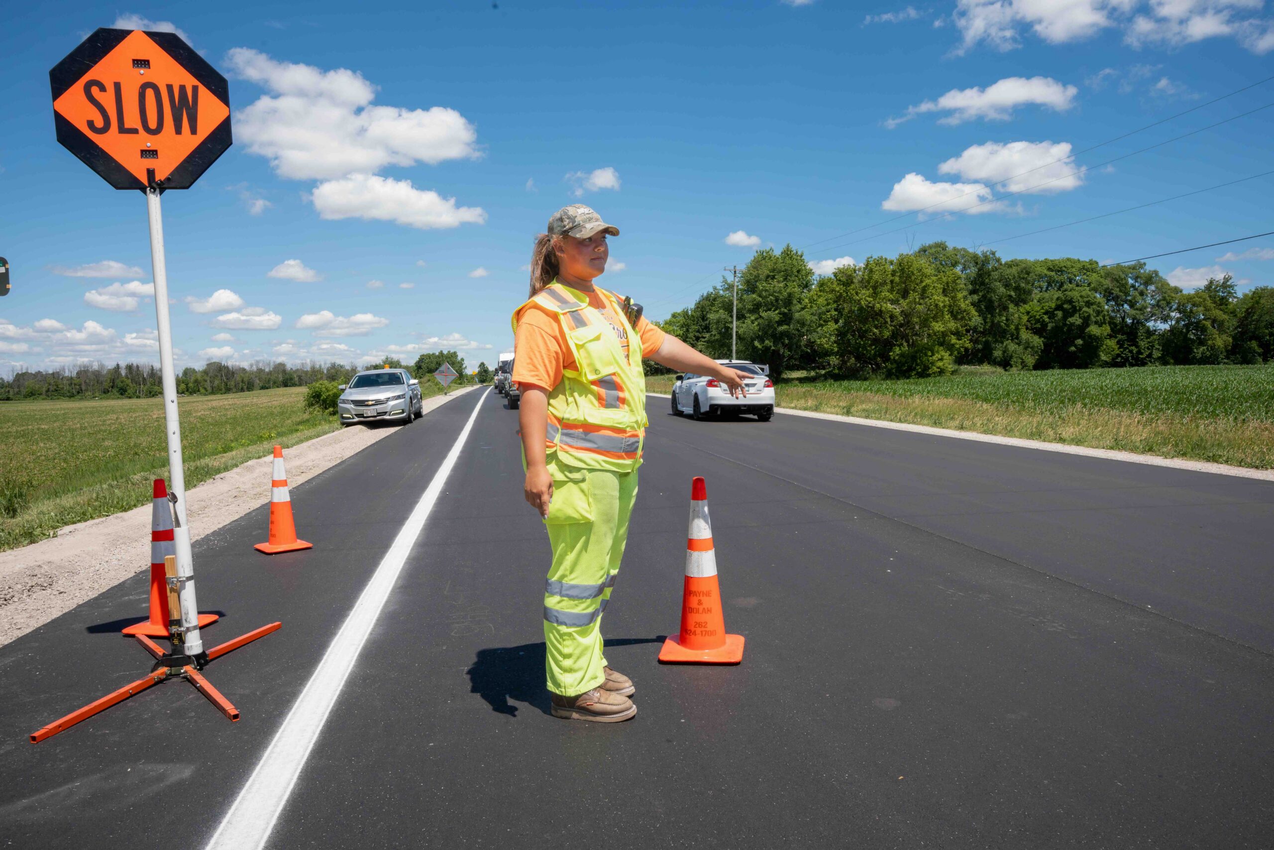 Road worker in high-visibility vest directing traffic beside a 'SLOW' sign and orange cones on a rural road.