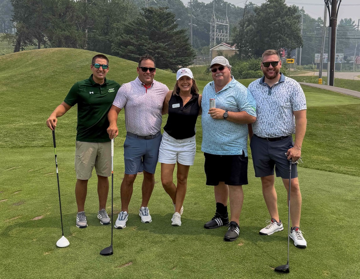 Five adults posing together on a golf green with clubs, smiling at the camera.
