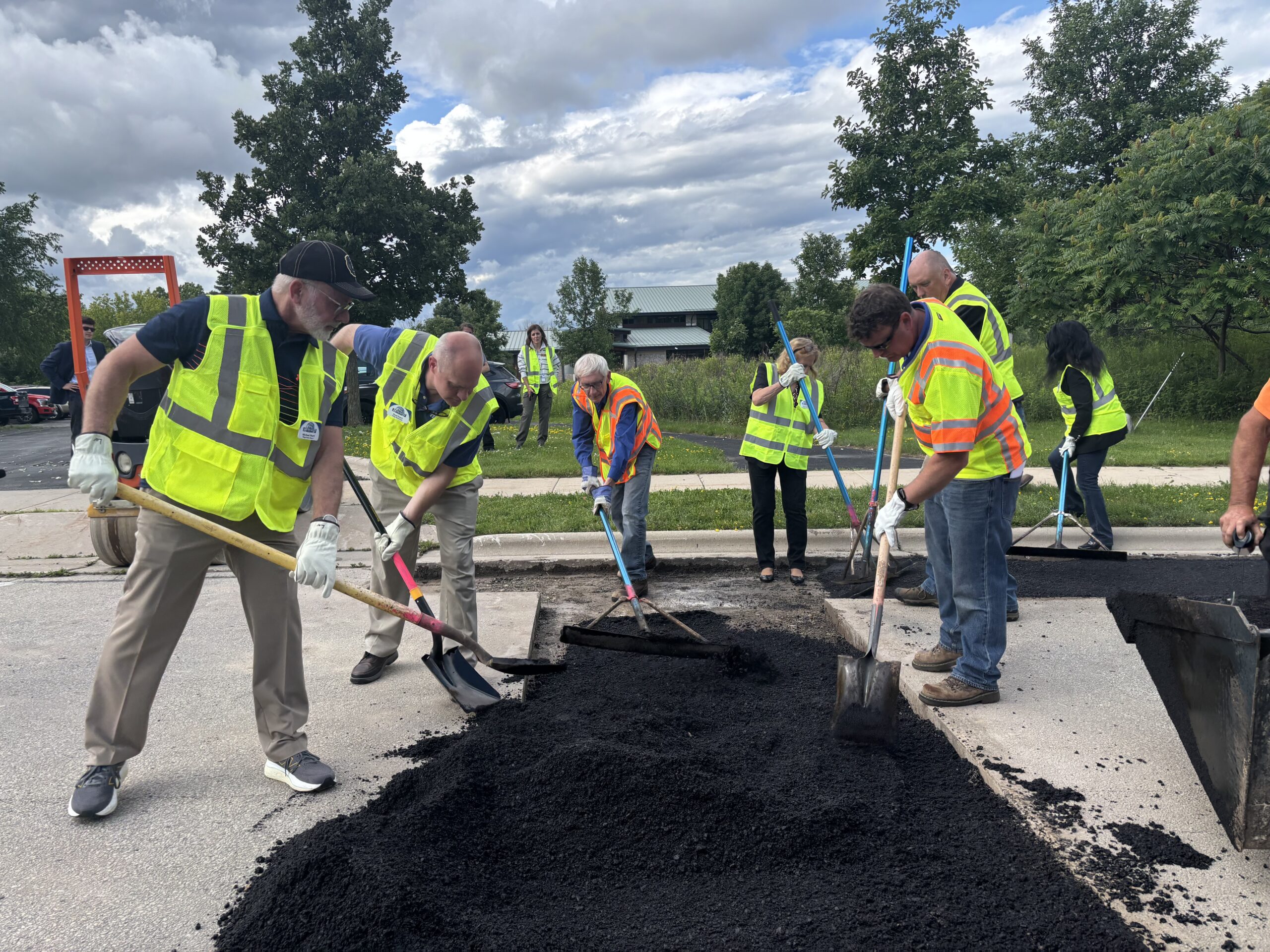 Construction workers in high‑visibility vests spread fresh asphalt on a road together.