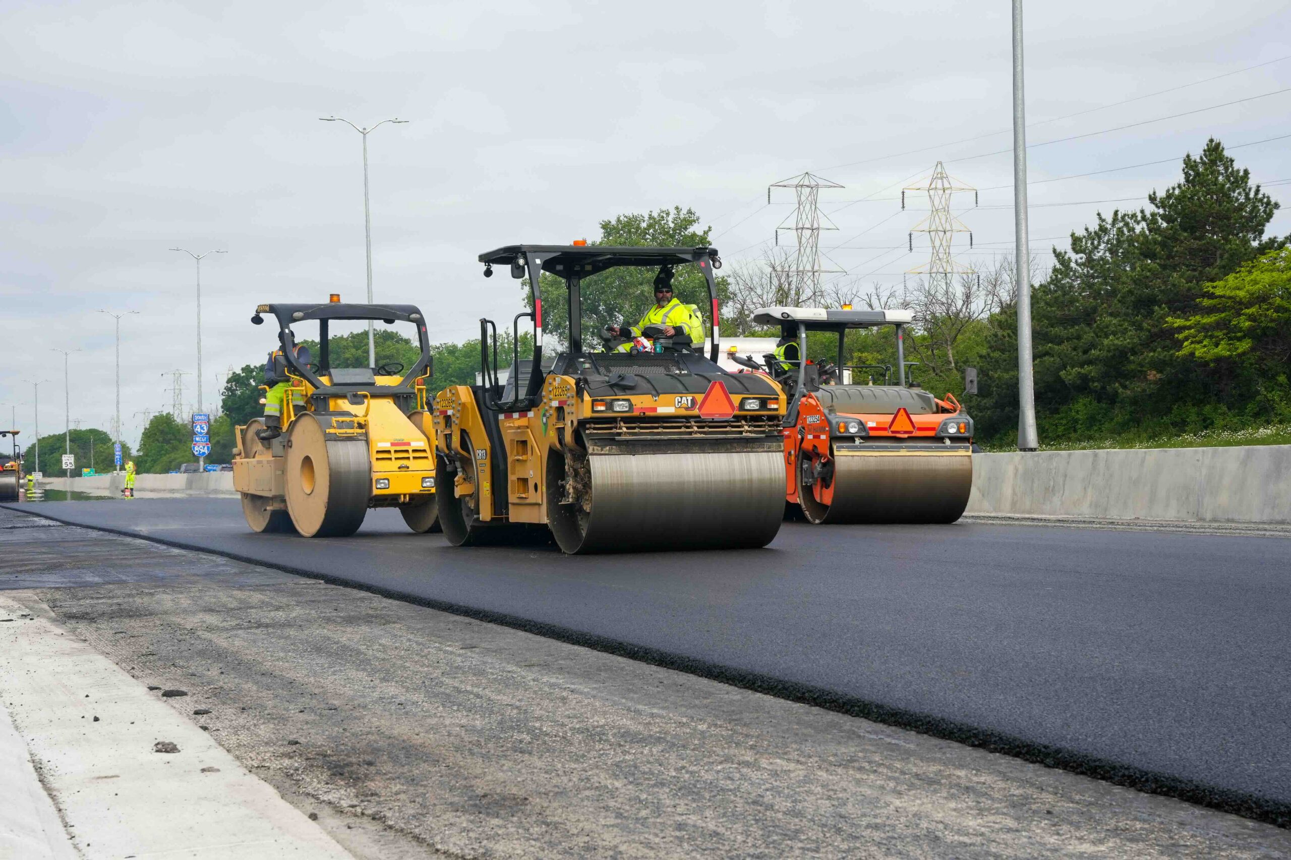 Road construction scene showing asphalt rollers compacting a highway, workers in hi-vis vests, and orange warning signs in the background.