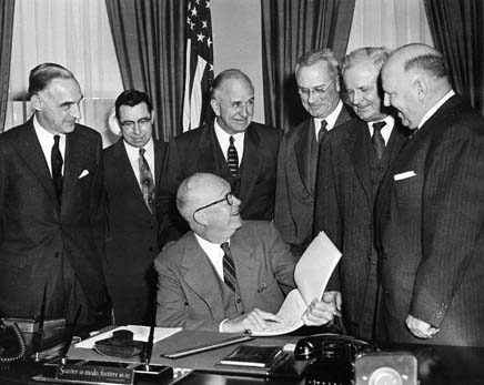 A man seated at a desk signs a document while seven suited men stand around, observing a formal signing ceremony.
