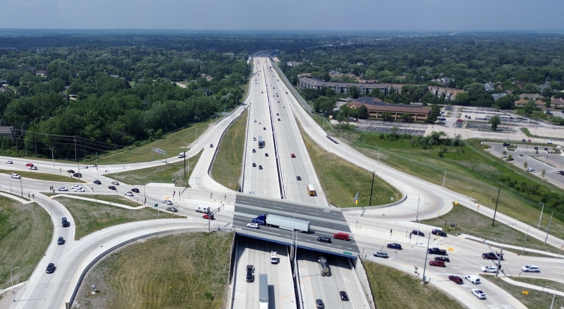 Aerial view of a multi-lane highway interchange surrounded by trees and suburban buildings, with multiple cars traveling in both directions on a sunny day far into the distance.