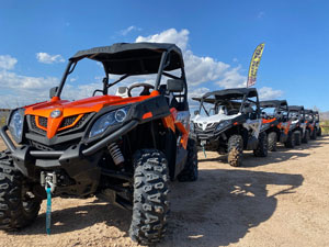 Row of orange and gray all-terrain vehicles (ATVs) parked on a dirt lot under a blue sky, ready for rental or tour.
