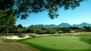 Golf course green with flagstick and sand bunker nearby, framed by trees and distant mountains on a sunny day.