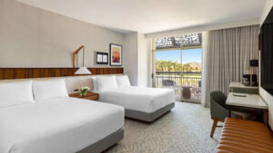 Hotel room with two queen beds, a shared nightstand, and a balcony visible through glass doors overlooking an outdoor seating area and sunny view.