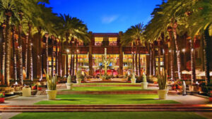 Resort courtyard at dusk with palm trees, a central fountain statue, and illuminated modern building in the background.