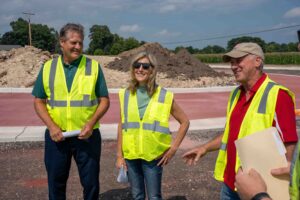 Three construction workers in high‑visibility vests stand at a dirt lot on a sunny day, looking off to the side and smiling. One holds papers.